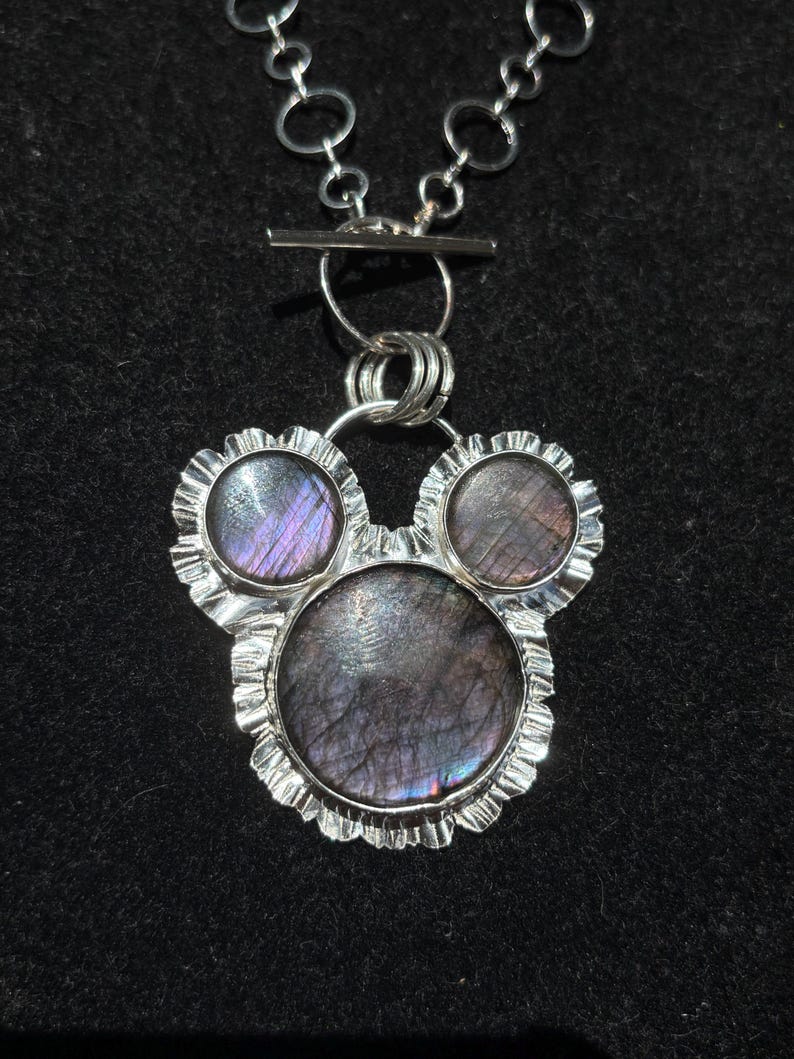 May include: A silver necklace featuring a pendant with three circular labradorite stones set in a ruffled metal frame. The stones display iridescent purple and grey tones. The pendant hangs from a silver chain with a toggle clasp closure.