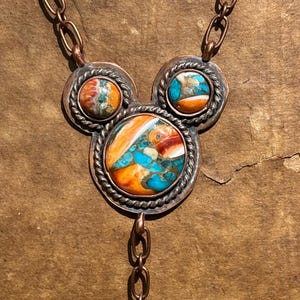 May include: A copper necklace featuring three circular settings arranged in a cluster. Each setting contains a multi-colored stone with swirls of orange, white, and turquoise blue. The stones are framed by twisted copper wire and linked by chains.