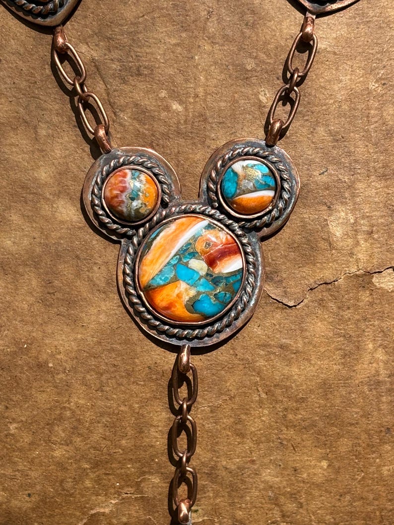 May include: A copper necklace featuring three circular settings arranged in a cluster. Each setting contains a multi-colored stone with swirls of orange, white, and turquoise blue. The stones are framed by twisted copper wire and linked by chains.