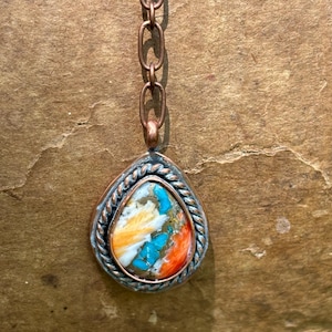 May include: A teardrop-shaped pendant featuring a multi-colored stone with streaks of white, orange, and bright turquoise, encased in a twisted metal border. The pendant hangs from a copper-toned chain with elongated oval links.
