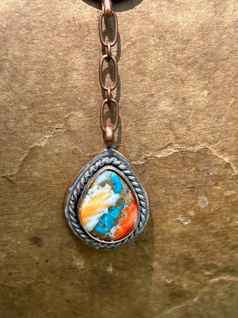 May include: A teardrop-shaped pendant featuring a multi-colored stone with streaks of white, orange, and bright turquoise, encased in a twisted metal border. The pendant hangs from a copper-toned chain with elongated oval links.