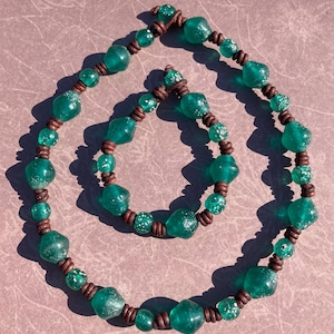 May include: A necklace and bracelet set featuring large, irregular, teal-colored glass beads strung on brown leather cord. The beads have a textured appearance, and the necklace and bracelet are displayed on a neutral surface.