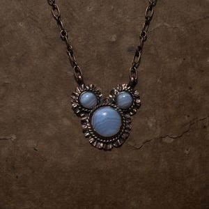 May include: A copper necklace featuring a pendant with three round, light blue banded agate stones set in scalloped metal frames. The central stone is larger than the two smaller stones above it. The pendant hangs from a matching copper chain link.