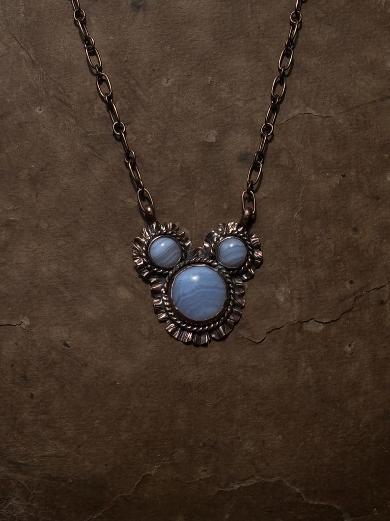 May include: A copper necklace featuring a pendant with three round, light blue banded agate stones set in scalloped metal frames. The central stone is larger than the two smaller stones above it. The pendant hangs from a matching copper chain link.