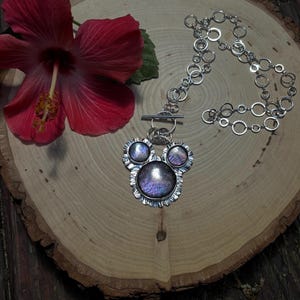 May include: A silver necklace featuring a pendant with three circular, iridescent purple stones set in scalloped metal frames. The pendant is attached to a chain made of interconnected silver rings and includes a horizontal bar toggle clasp.