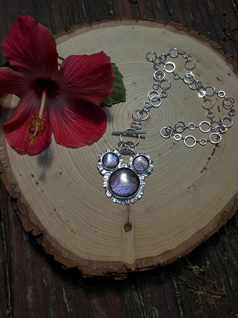 May include: A silver necklace featuring a pendant with three circular, iridescent purple stones set in scalloped metal frames. The pendant is attached to a chain made of interconnected silver rings and includes a horizontal bar toggle clasp.