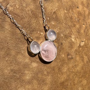 Puede incluir: Un collar de plata con tres piedras de cuarzo rosa. El collar tiene una cadena fina y las piedras est&aacute;n engastadas en biseles de plata. La piedra m&aacute;s grande es redonda y de color rosa claro, las otras dos son m&aacute;s peque&ntilde;as y claras.