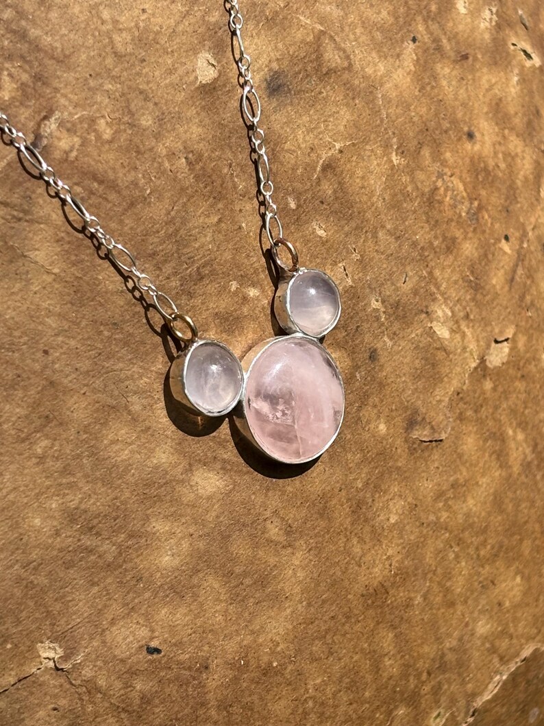 Puede incluir: Un collar de plata con tres piedras de cuarzo rosa. El collar tiene una cadena fina y las piedras est&aacute;n engastadas en biseles de plata. La piedra m&aacute;s grande es redonda y de color rosa claro, las otras dos son m&aacute;s peque&ntilde;as y claras.