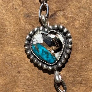 May include: A heart-shaped pendant featuring a mosaic of turquoise, white, and black stone segments. The pendant is framed by a beaded silver border and is attached to a silver chain with oval links extending from the top and bottom.