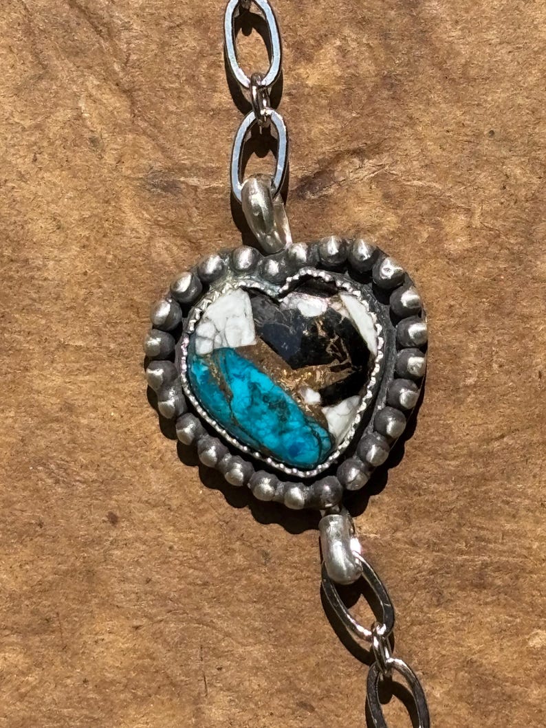May include: A heart-shaped pendant featuring a mosaic of turquoise, white, and black stone segments. The pendant is framed by a beaded silver border and is attached to a silver chain with oval links extending from the top and bottom.