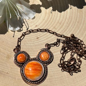 May include: A copper-toned necklace with a pendant featuring three orange and white spiny oyster shell stones. The necklace has a chain and is displayed on a wooden surface, with white flowers in the background.