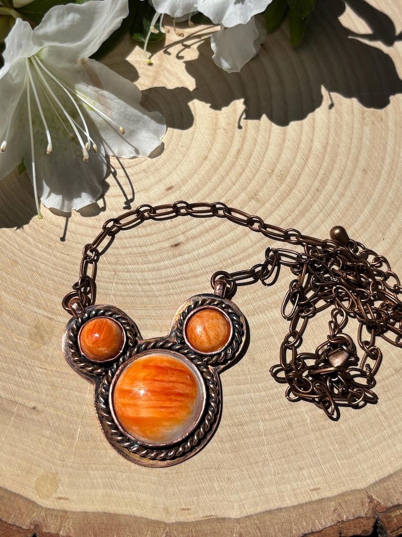 May include: A copper-toned necklace with a pendant featuring three orange and white spiny oyster shell stones. The necklace has a chain and is displayed on a wooden surface, with white flowers in the background.