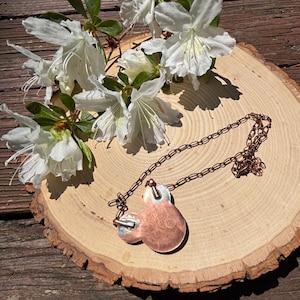 May include: A copper pendant necklace with a unique design, resting on a wooden slice. The necklace features a chain and a pendant with a circular shape. White flowers with green leaves are arranged around the necklace.