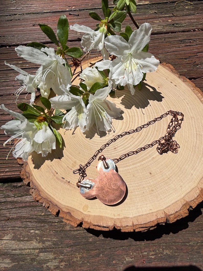 May include: A copper pendant necklace with a unique design, resting on a wooden slice. The necklace features a chain and a pendant with a circular shape. White flowers with green leaves are arranged around the necklace.