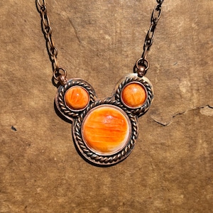 May include: A copper-toned necklace featuring three orange-colored, round stones. The stones are set in a decorative frame with a twisted rope design. The necklace has a matching copper chain.