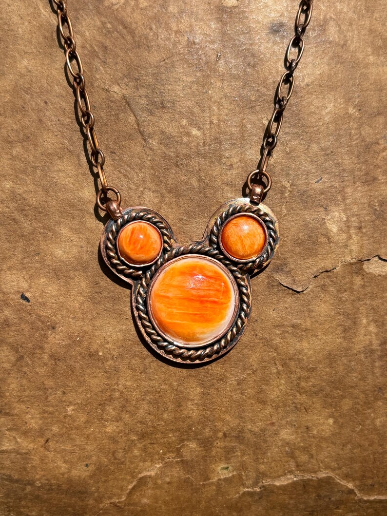 May include: A copper-toned necklace featuring three orange-colored, round stones. The stones are set in a decorative frame with a twisted rope design. The necklace has a matching copper chain.