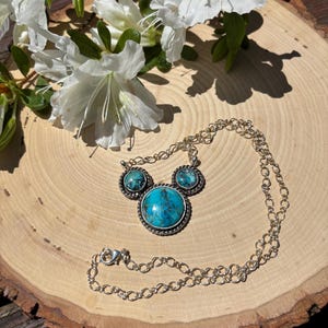 Pu&ograve; includere: Una collana d'argento con una grande pietra turchese rotonda al centro, affiancata da due pietre turchesi pi&ugrave; piccole. La collana &egrave; su una superficie di legno, con fiori bianchi sullo sfondo.