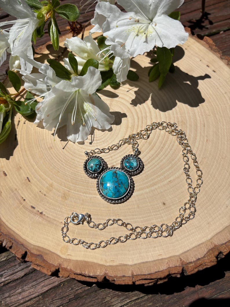 Pu&ograve; includere: Una collana d'argento con una grande pietra turchese rotonda al centro, affiancata da due pietre turchesi pi&ugrave; piccole. La collana &egrave; su una superficie di legno, con fiori bianchi sullo sfondo.