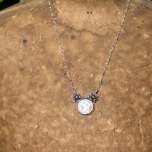 K&ouml;nnte beinhalten: Eine silberfarbene Halskette mit einem runden Anh&auml;nger in der Mitte, der ein Reliefdesign aufweist und von zwei kleinen blumenf&ouml;rmigen Akzenten flankiert wird. Der Anh&auml;nger h&auml;ngt an einer zarten, d&uuml;nnen Metallkette mit l&auml;nglichen ovalen Gliedern.