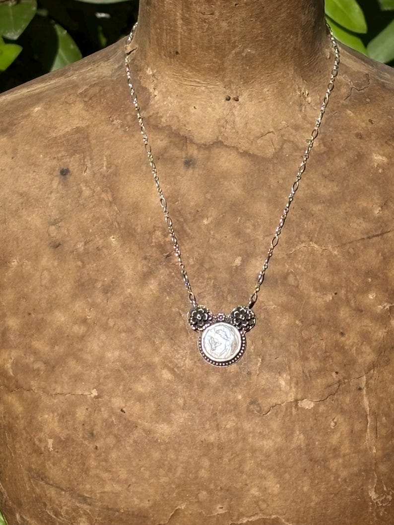 K&ouml;nnte beinhalten: Eine silberfarbene Halskette mit einem runden Anh&auml;nger in der Mitte, der ein Reliefdesign aufweist und von zwei kleinen blumenf&ouml;rmigen Akzenten flankiert wird. Der Anh&auml;nger h&auml;ngt an einer zarten, d&uuml;nnen Metallkette mit l&auml;nglichen ovalen Gliedern.