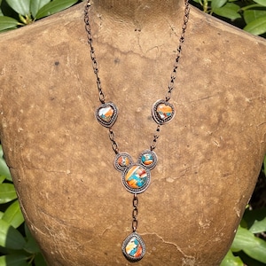 May include: A copper chain necklace featuring five inlaid stone settings with vibrant orange, blue, and white marbled patterns. The design includes two heart-shaped stones, two small circular stones, one large central circular stone, and a teardrop pendant.