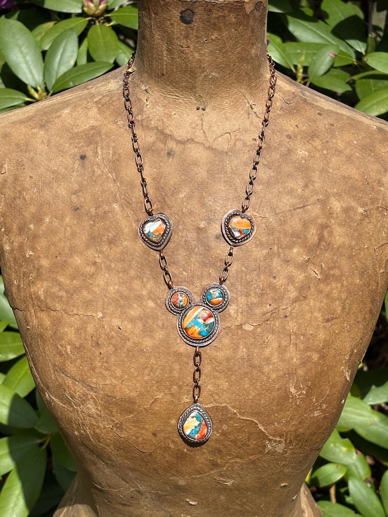 May include: A copper chain necklace featuring five inlaid stone settings with vibrant orange, blue, and white marbled patterns. The design includes two heart-shaped stones, two small circular stones, one large central circular stone, and a teardrop pendant.