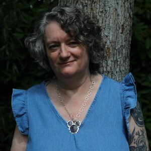 May include: A silver-toned necklace featuring a chain of interconnected circular links and a central pendant with three dark, round stones set in a decorative silver frame, worn with a blue sleeveless top with ruffled shoulder details.