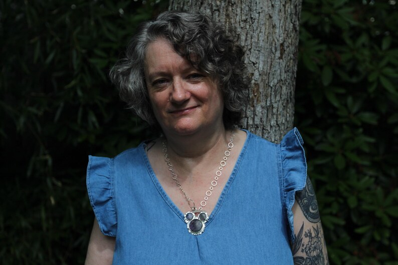May include: A silver-toned necklace featuring a chain of interconnected circular links and a central pendant with three dark, round stones set in a decorative silver frame, worn with a blue sleeveless top with ruffled shoulder details.