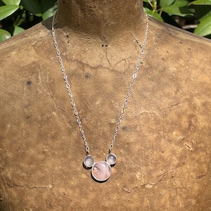 Puede incluir: Un collar de plata con tres piedras de cuarzo rosa. La piedra central es de forma redonda y m&aacute;s grande, flanqueada por dos piedras circulares m&aacute;s peque&ntilde;as. La cadena tiene un dise&ntilde;o de eslabones delicados. El collar se exhibe en un maniqu&iacute;.