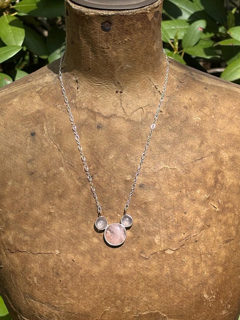 Puede incluir: Un collar de plata con tres piedras de cuarzo rosa. La piedra central es de forma redonda y m&aacute;s grande, flanqueada por dos piedras circulares m&aacute;s peque&ntilde;as. La cadena tiene un dise&ntilde;o de eslabones delicados. El collar se exhibe en un maniqu&iacute;.
