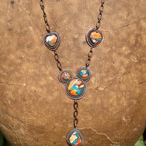 May include: A copper chain necklace featuring five inlaid stone settings with vibrant orange, turquoise, and white marbled patterns. The design includes two heart-shaped accents, two circular accents, and a central teardrop-shaped pendant.