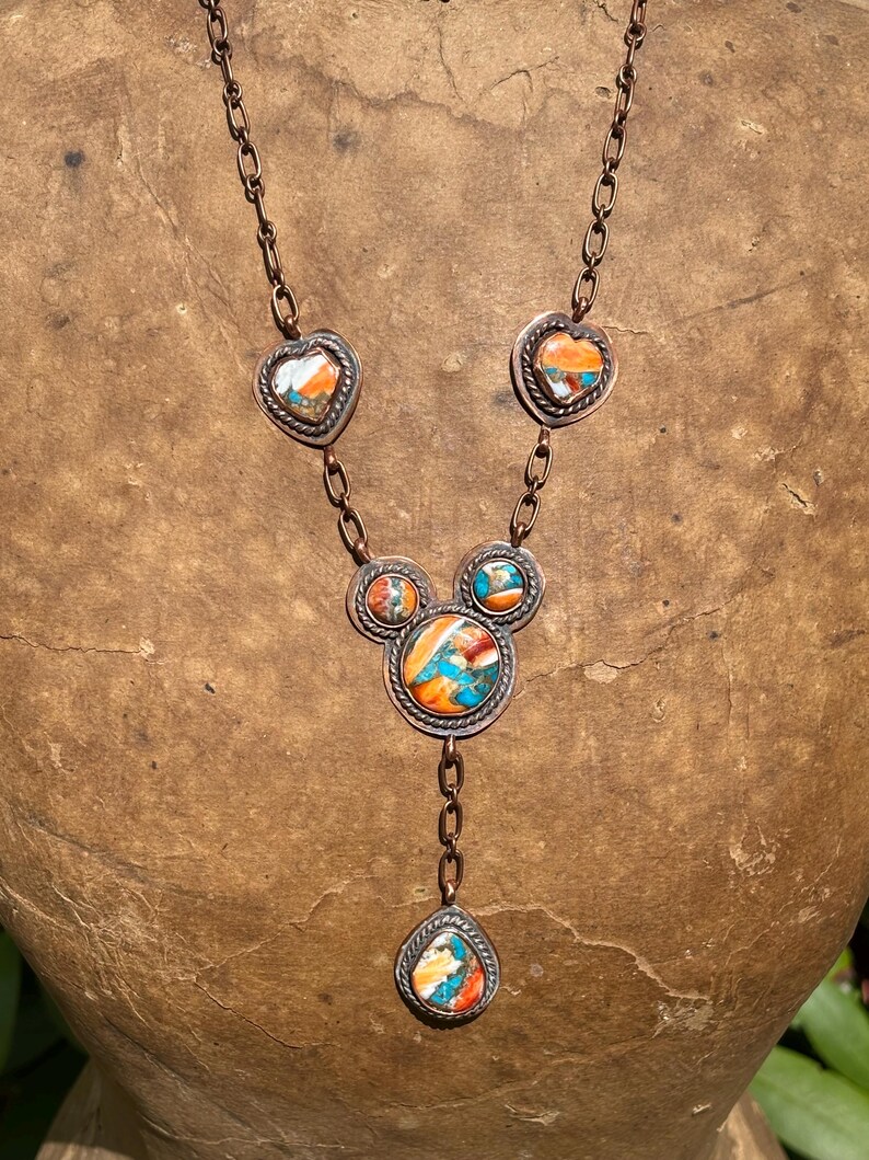 May include: A copper chain necklace featuring five inlaid stone settings with vibrant orange, turquoise, and white marbled patterns. The design includes two heart-shaped accents, two circular accents, and a central teardrop-shaped pendant.