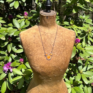 May include: A copper-toned necklace with three orange-colored circular pendants is displayed on a vintage dress form. The necklace is suspended from a delicate chain. The dress form is set against a backdrop of green foliage and purple flowers.