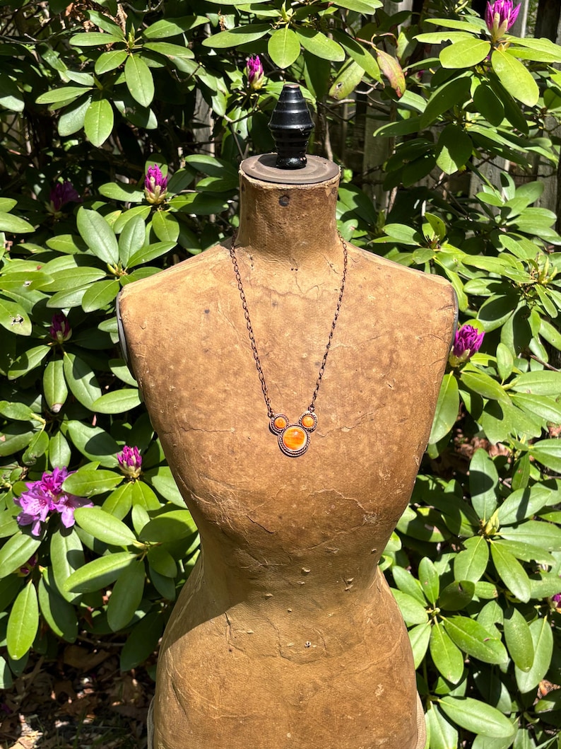 May include: A copper-toned necklace with three orange-colored circular pendants is displayed on a vintage dress form. The necklace is suspended from a delicate chain. The dress form is set against a backdrop of green foliage and purple flowers.
