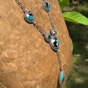 May include: A silver necklace featuring a series of linked metal settings containing inlaid stones with black and turquoise blue patterns. The design includes heart-shaped and circular elements with beaded edges, connected by a delicate silver chain.