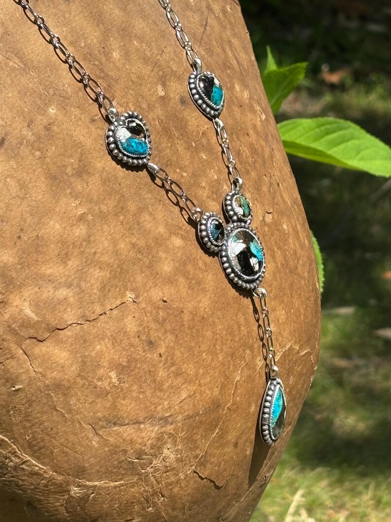 May include: A silver necklace featuring a series of linked metal settings containing inlaid stones with black and turquoise blue patterns. The design includes heart-shaped and circular elements with beaded edges, connected by a delicate silver chain.