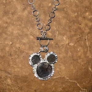May include: A silver necklace featuring a chain of circular links and a toggle clasp. The pendant consists of three dark, iridescent circular stones set in silver with a ruffled, textured edge, arranged in a cluster resembling a rounded shape.