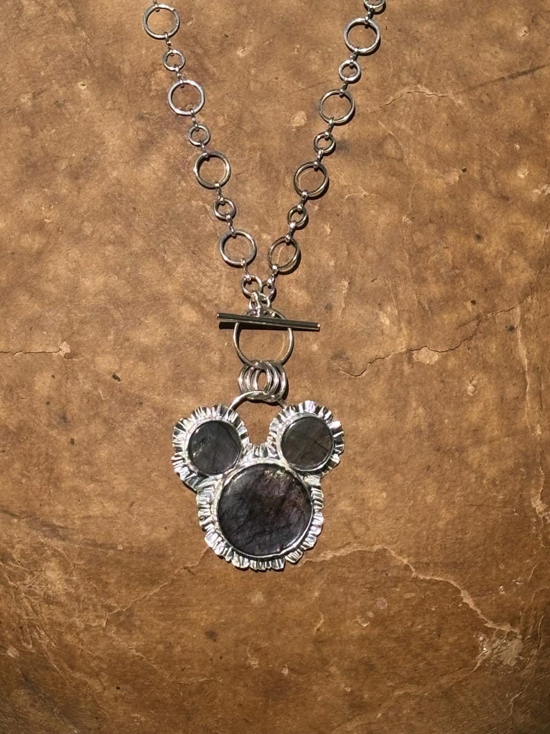 May include: A silver necklace featuring a chain of circular links and a toggle clasp. The pendant consists of three dark, iridescent circular stones set in silver with a ruffled, textured edge, arranged in a cluster resembling a rounded shape.
