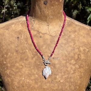 May include: A necklace featuring a strand of small, deep pink beads. The necklace has a silver clasp and a large, irregularly shaped white pearl pendant. The necklace is displayed on a mannequin.