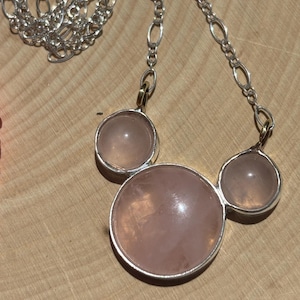 Puede incluir: Un collar de plata con tres piedras redondas de cuarzo rosa p&aacute;lido. Las piedras est&aacute;n engastadas en biseles de plata y conectadas por una cadena de plata. El collar se muestra sobre una superficie de madera.