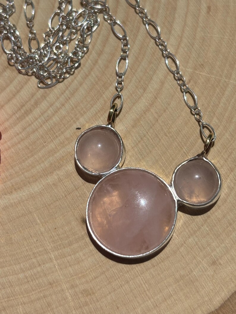 Puede incluir: Un collar de plata con tres piedras redondas de cuarzo rosa p&aacute;lido. Las piedras est&aacute;n engastadas en biseles de plata y conectadas por una cadena de plata. El collar se muestra sobre una superficie de madera.
