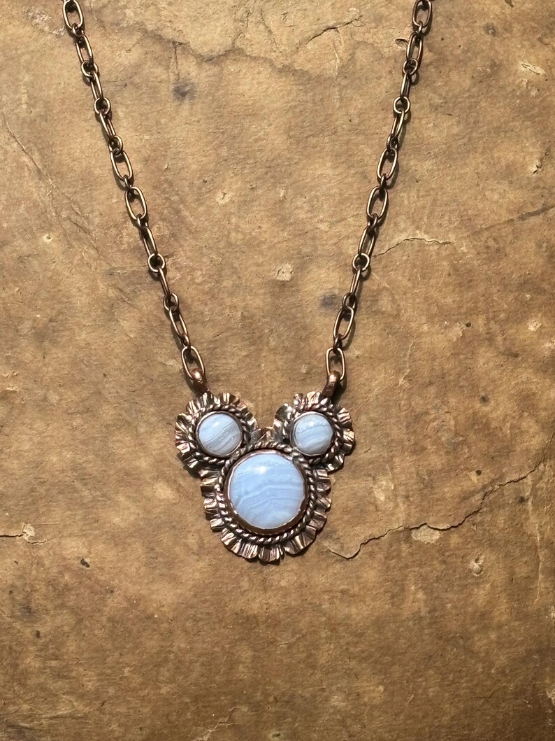 May include: A necklace featuring a copper chain and a pendant with three round, light blue, banded agate stones set in copper. The stones are arranged in a cluster, with one large stone at the bottom and two smaller stones above, all framed by textured copper metalwork.