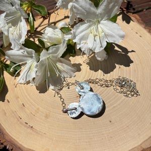 Pu&ograve; includere: Una collana con pendente argentato a forma di testa di topo, con una catena argentata. Il pendente &egrave; su una superficie di legno, con fiori bianchi e foglie verdi sullo sfondo.