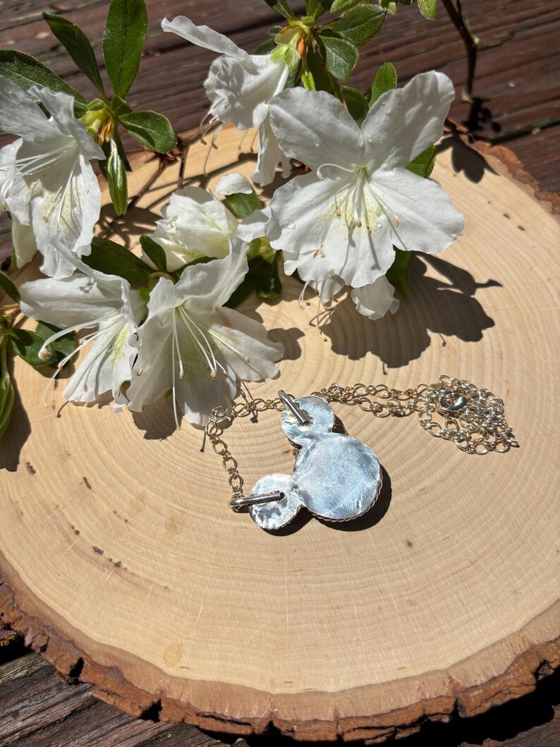 Pu&ograve; includere: Una collana con pendente argentato a forma di testa di topo, con una catena argentata. Il pendente &egrave; su una superficie di legno, con fiori bianchi e foglie verdi sullo sfondo.