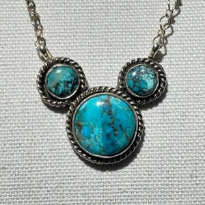 Pu&ograve; includere: Una collana color argento con tre pietre turchesi. Le pietre sono incastonate in un bordo argentato a forma di corda. La pietra centrale &egrave; pi&ugrave; grande delle due laterali. Anche la catena della collana &egrave; argentata.