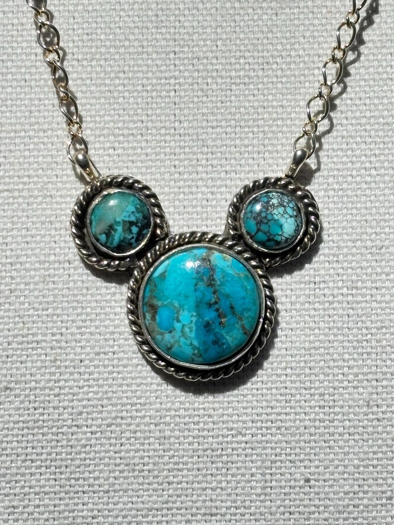 Pu&ograve; includere: Una collana color argento con tre pietre turchesi. Le pietre sono incastonate in un bordo argentato a forma di corda. La pietra centrale &egrave; pi&ugrave; grande delle due laterali. Anche la catena della collana &egrave; argentata.