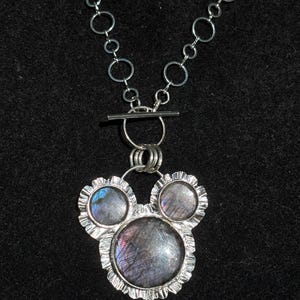 May include: A silver-toned necklace featuring a pendant with three circular labradorite stones set in scalloped metal frames. The pendant is suspended from a chain composed of interconnected silver rings and includes a horizontal bar toggle clasp.