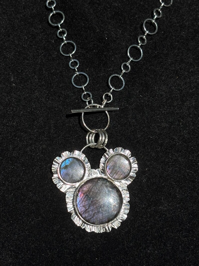 May include: A silver-toned necklace featuring a pendant with three circular labradorite stones set in scalloped metal frames. The pendant is suspended from a chain composed of interconnected silver rings and includes a horizontal bar toggle clasp.