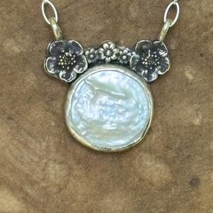 May include: A necklace featuring a round, iridescent white coin pearl pendant set in a metallic frame. The pendant is topped with a dark, oxidized silver floral motif consisting of two larger flowers and a smaller central blossom, attached to a silver chain.