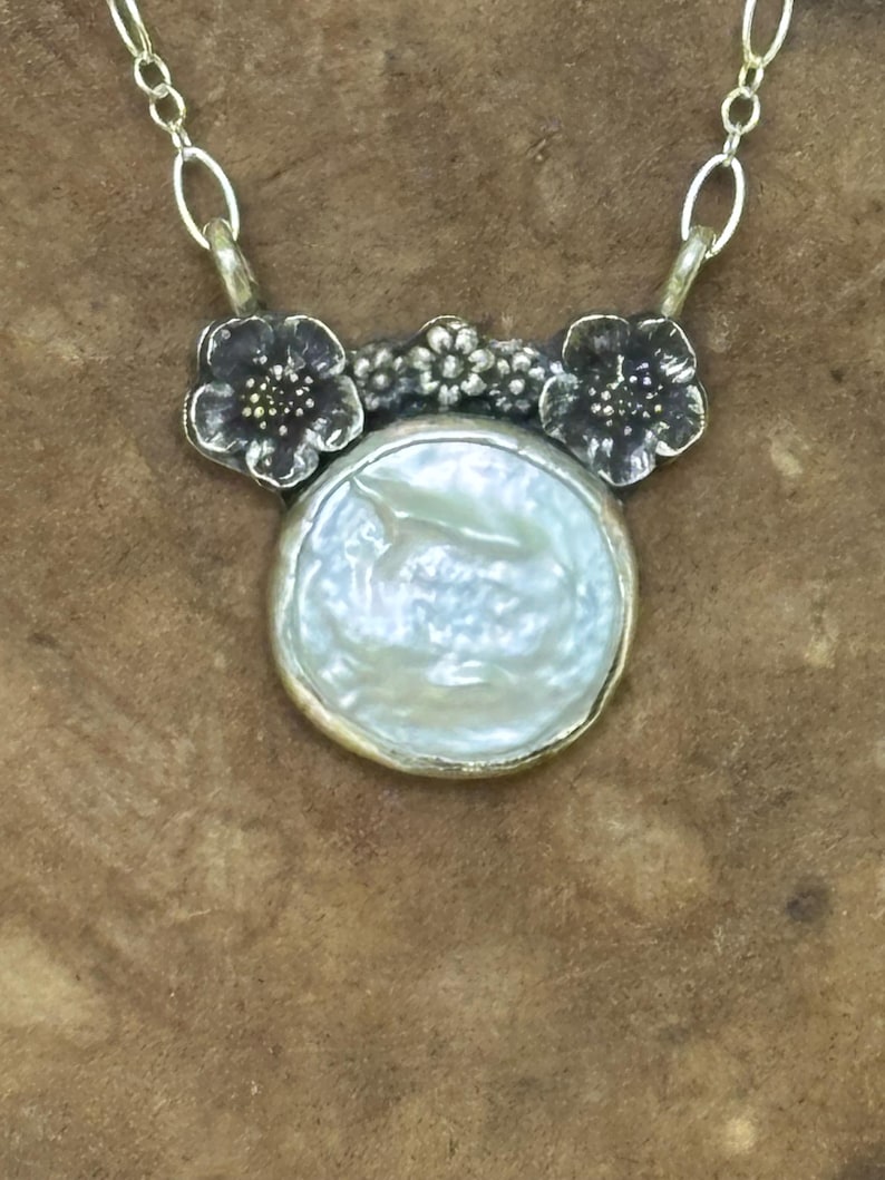 May include: A necklace featuring a round, iridescent white coin pearl pendant set in a metallic frame. The pendant is topped with a dark, oxidized silver floral motif consisting of two larger flowers and a smaller central blossom, attached to a silver chain.